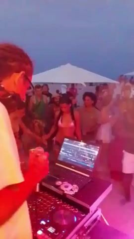 The video shows a crowded beach party at dusk. A DJ, wearing headphones and holding a microphone, is positioned in the foreground, operating a laptop and DJ equipment. A diverse crowd of people is dancing and socializing behind the DJ. The environment features white umbrellas and a hazy, blue-toned sky, indicating the late hour. The lighting is dynamic, with pink and purple hues illuminating the scene.
