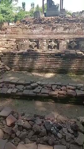 The video pans across a section of ancient ruins made of brick and stone. In the foreground, there are piles of rubble and broken bricks, suggesting a state of disrepair. The ruins appear to be tiered, with visible brick walls and stone foundations. In the background, a simple wooden structure with a peaked roof covers a large stone statue or monument. Beyond the ruins, there are trees, vegetation, and some modern buildings, indicating the site is likely located in a populated area. The sky is visible and appears to be clear and sunny. The camera movement is slow and deliberate, allowing for a detailed observation of the archaeological site. There are no people visible in the video.