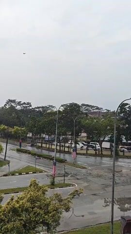 A cloudy day with light rain falls on a wet intersection of roads. In the foreground, a few cars and a motorcycle drive through the intersection. The intersection is marked with white lines and a traffic signal light is visible. In the background, there are trees and some buildings. Malaysian flags are displayed on poles along the road. The overall scene is peaceful and serene, with the rain creating a soft, diffused light.