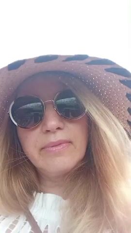 A woman with blonde hair, wearing a wide-brimmed hat with a leopard-print band and round sunglasses, is looking at the camera. She has on a white top with decorative cutouts. Her mouth is slightly open. The video appears to be a selfie taken outdoors, as suggested by the bright lighting and the reflection of what looks like buildings or trees in her sunglasses.