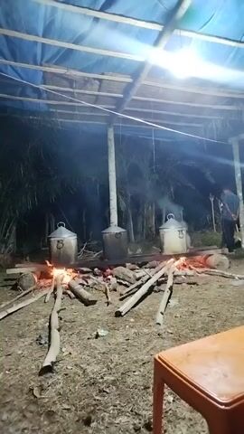 The video shows an outdoor cooking scene at night. Three large, silver pots are placed over open fires. The pots appear to be cooking something, as evidenced by the steam rising from them. The fires are made of logs and branches. The scene is lit by a single overhead light, casting long shadows. In the background, there are trees and foliage. A person can be seen standing to the right of the frame, partially obscured by darkness. A wooden table is visible in the foreground.