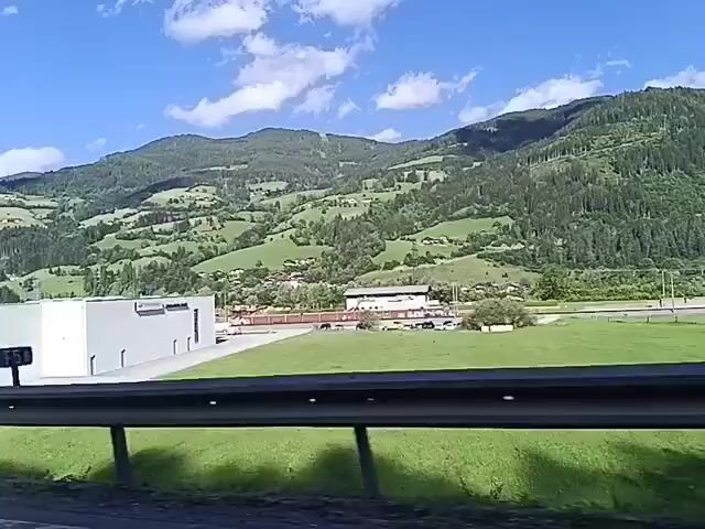 The video shows a roadside view of a rural mountainous area with green hills and trees under a blue sky with some clouds. There are buildings and a parking lot with cars visible in the middle ground. The foreground features a black metal guardrail. The video appears to be recorded from a moving vehicle, as evidenced by the slight camera shake and the changing perspective.