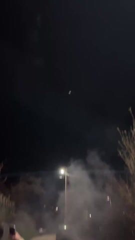 The video shows fireworks going off in the night sky, followed by a quick pan to a group of people holding a banner. The fireworks are red, white, and yellow. The banner is a white cloth covered with graffiti. The people holding the banner are mostly men and wearing casual clothing. They are standing on a brick sidewalk. There is a grassy area and some trees in the background. The environment is dark, and there is some light coming from streetlights.