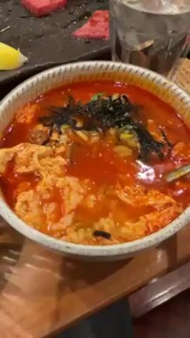 The video shows a person stirring a bowl of what appears to be a spicy rice soup or congee with a spoon. The soup has an orange-red color and contains rice, shredded egg, and seaweed. A glass of water sits in the background, along with a black plate holding pieces of red meat and a slice of lemon. The bowl rests on a wooden surface.