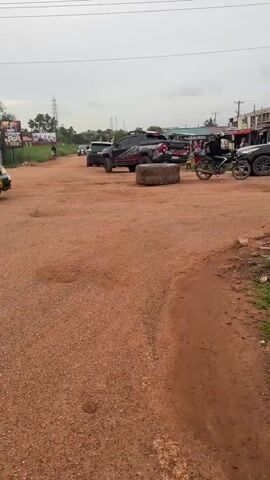 The video depicts a red dirt road in what appears to be a residential or commercial area with various buildings and signs visible in the background. The weather appears to be overcast.  Several vehicles are present, including a taxi, a pickup truck, and an SUV. The road is partially blocked by a large stone. The camera moves to the right, revealing a roadside stall where men are preparing coconuts. One man is shirtless. The stall has a sign that reads "Evans Co Fresh Coconut." There is also some trash, including discarded tires and other debris.