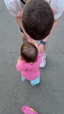 A top-down medium shot shows a man in a white t-shirt and black shorts interacting with a young child wearing a pink t-shirt and blue striped shorts. The man is leaning down and appears to be wiping something off the child's hand with a white tissue. The child is standing, facing away from the camera, with their back to the man. The ground beneath them is asphalt. The man has a dark wristwatch on his left wrist and a tattoo on his left calf. The child has dark hair and is wearing blue sneakers. Another child's pink sneaker with yellow laces is visible on the ground in the lower foreground.