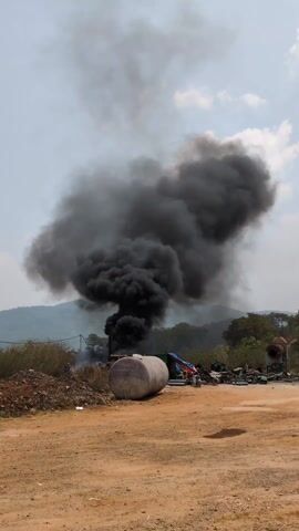 In the video, a large plume of black smoke rises from an area with industrial debris. The smoke is thick and dark, contrasting against the partly cloudy sky. In the foreground, there's a cylindrical tank lying on its side on a dirt ground, with piles of debris nearby. Behind the tank, the source of the smoke appears to be a structure obscured by the smoke itself. The background shows a landscape with trees and hills. The environment looks like an industrial or construction site. There are no visible people or specific actions beyond the smoke billowing.