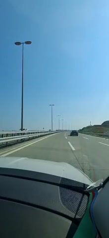 This is a first-person perspective video taken from inside a car traveling on a highway. The highway appears to be modern and well-maintained, with multiple lanes and a clear blue sky overhead, suggesting a bright, sunny day. The road is bordered by a metal guardrail on the left, with what looks like a body of water or a large flat expanse in the distance beyond it. On the right side of the highway, a gentle slope covered in dry vegetation rises upwards.

The primary activity is driving on the highway. Several other vehicles are visible: a dark-colored SUV is in front and to the right of the camera's car, and further ahead, other cars can be seen on the road. The camera car itself has a white hood and dashboard visible in the foreground. The clear sky and the dry vegetation hint at a warm climate, possibly late spring or summer.

Notable details include the presence of numerous tall lampposts lining the highway, suggesting it's designed for both day and night use. The clear, unblemished blue sky and the sunlight casting a slight glare on the windshield indicate a sunny day with no clouds. The video's timestamp shows it was recorded around midday. There is no visible text within the media itself. The overall mood conveyed is one of smooth travel on an open road under pleasant weather conditions. The location context of Surakhany Raion, Azerbaijan, helps to place this highway within a specific geographical region.