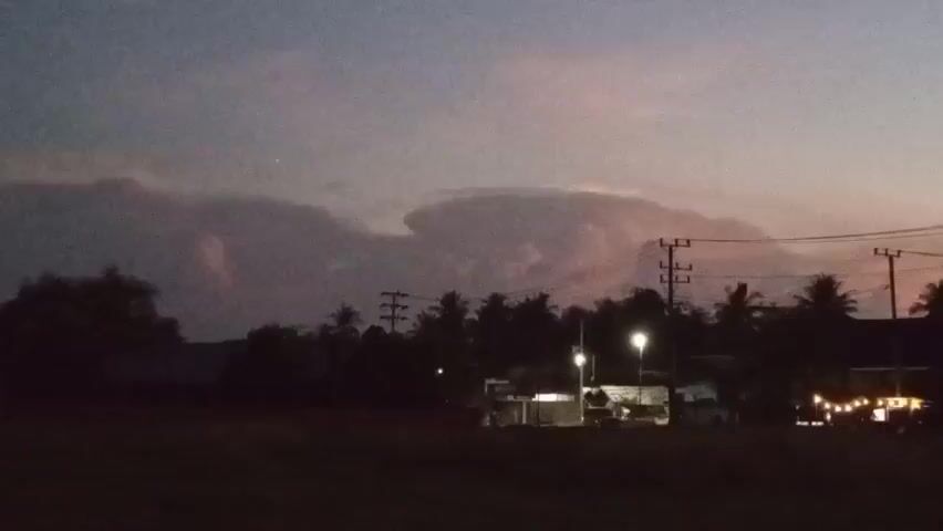 A time-lapse video captures the twilight sky above a silhouetted landscape. The sky transitions from a deep purple-blue to shades of pink and orange as the sun sets. Dominating the sky is a large, unusual cloud formation that resembles a lenticular cloud, with a distinct saucer-like shape. Below the sky, a dark silhouette of trees and palm trees lines the horizon. In the mid-ground, there are faint outlines of buildings and structures, illuminated by artificial lights, including streetlights and string lights on what appears to be a building on the right side of the frame. Electrical poles and wires are visible, adding to the rural or semi-rural setting. The foreground is dark, obscuring any details on the ground. The video appears to be taken from a stationary viewpoint, observing the slow changes in the sky and ambient lighting.