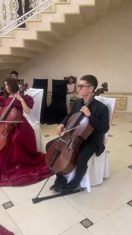 The video shows a string quartet performing indoors in a well-lit, ornate room with a grand staircase in the background. Three musicians are clearly visible: a man and two women, all dressed in elegant, floor-length, deep red gowns. The man, on the right, is playing a cello. The woman next to him, in the center, is also playing a cello. The woman on the left is playing a violin. Their instruments are propped on stands. There are black chairs behind them, and a young girl can be seen sitting behind the chairs. The floor is tiled with a patterned design. The musicians are seated and appear to be focused on their performance. The lighting is warm and bright, illuminating the gold accents on the staircase and the walls. The overall atmosphere suggests a formal event or concert.