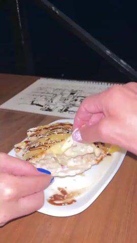 A close-up shot shows a person's hands separating a stuffed flatbread or pastry on a white rectangular plate. The filling appears to be cheese and possibly meat, and it is gooey and stretchy as the pastry is pulled apart. The pastry itself is golden brown and appears to be drizzled with a dark sauce and sprinkled with sesame seeds. A lemon wedge is visible on the side of the plate. In the background, a menu with an illustration is partially visible on a wooden table. The lighting is dim, creating a cozy atmosphere.