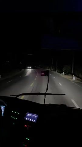 The video provides a first-person perspective from inside a vehicle at night. The headlights of the vehicle illuminate a dark, paved road with yellow and white lane markings. The road appears to be a two-lane highway, with a shoulder on the right side bordered by sparse vegetation and utility poles. In the distance, two sets of headlights from other vehicles are visible on the road ahead. The interior of the vehicle is dimly lit, with glowing green and red indicator lights visible on the dashboard. A windshield wiper is visible in the foreground, and a navigation or infotainment screen is also apparent on the dashboard. The overall impression is of driving at night on an open road.