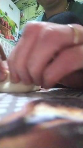 A woman is sitting at a table and working with a piece of white dough. She uses her fingers to pinch and shape the dough into a sort of cup-like form. She's wearing a green sweater and black leggings. Behind her is a refrigerator decorated with images of fruit and a calendar. The table is covered with a patterned tablecloth. The focus is on her hands as she manipulates the dough.