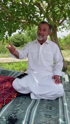 An eye-level shot shows a man sitting under a tree being recorded on a smartphone. The man, who has a graying beard and mustache, is wearing a white robe. He sits cross-legged on a striped mat with a red patterned blanket underneath. A black smartphone sits next to him on the mat. The person holding the smartphone is wearing a short-sleeved shirt. The background is green and lush, with trees and grass visible. The man is gesturing with his hands as he speaks to the person filming.
