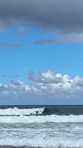 A man is surfing on a wave in the ocean. He is wearing a red shirt. The sky is partly cloudy with a mix of blue sky and grey storm clouds. The waves are moderate in size. The water is dark. The camera is stationary.