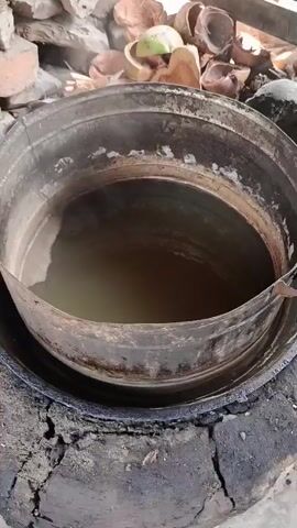 This video captures a close-up view of a rustic setup for boiling liquid. A large, weathered metal pot sits within a makeshift furnace, likely made of mud and rock. The pot contains a murky, yellowish liquid that is visibly steaming, indicating it is hot. The furnace itself appears to be constructed from a large, hollowed-out log, with openings for fuel and airflow. To the left of the pot, several pieces of firewood are visible, some charred from previous use. The background is filled with a large pile of cut logs and some dried coconut shells, suggesting that these materials are used as fuel for the fire. The overall environment appears to be outdoors, possibly in a rural or traditional setting, with a worn and functional aesthetic. The lighting is natural, and the camera movement is steady, focusing on the boiling pot and its immediate surroundings.