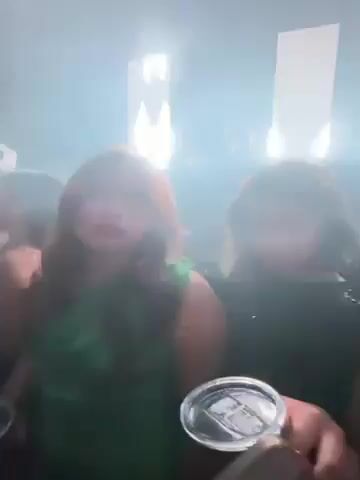 The video shows a group of young women at what appears to be a dance club or concert. They are taking a selfie video with a phone. The lighting changes colors frequently, including red, white, and blue. There are other people in the background and a DJ can be seen on a raised platform with rectangular lights behind them. The music is loud.
