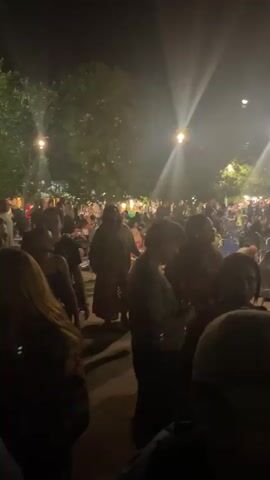 The video shows a crowded outdoor event at night, illuminated by bright spotlights. The camera moves through the crowd towards a stage where a performance seems to be taking place. People are gathered, some seated and some standing. The lighting suggests a festive atmosphere. The exact nature of the event is unclear from the visuals alone.