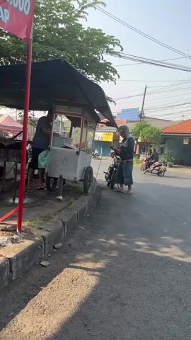 The video shows a street scene with a food cart. Two men are standing near the food cart. One man is wearing a dark shirt and shorts and is looking down at something on the cart. The other man is wearing a helmet and a blue shirt and is standing next to a motorcycle. The food cart is under a black tarp. There is a red sign on a pole to the left of the cart. The ground is asphalt and there is a sidewalk. In the background, there are houses and trees. A motorcycle is also visible in the background. The sun is shining, casting shadows.