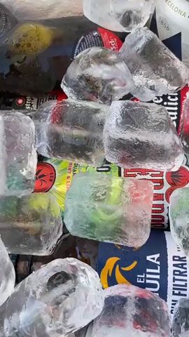 The video shows a close-up view of a cooler filled with ice and beverages. Several bottles and cans are visible, mostly obscured by large chunks of ice. Some of the beverages appear to be beer bottles, with labels indicating they are from different brands. The ice is clear and some pieces are tinged with color from the labels of the beverages. The overall scene suggests a refreshing set-up for drinks in a warm environment. In the last few seconds, a hand is seen placing a plastic bag over some of the ice.
