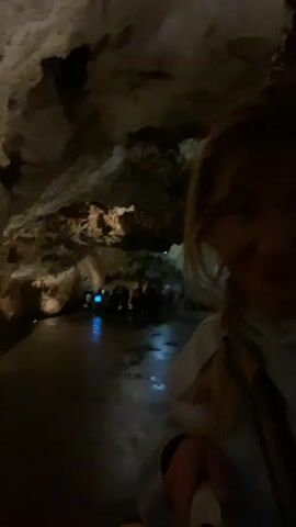 A woman is taking a selfie video inside a cave. She is wearing a light blue hooded sweatshirt and has blonde hair styled in a braid. The cave is dimly lit, with the walls and ceiling appearing rugged and textured. In the background, a path or walkway is visible with dim lighting illuminating the way. Other people are also walking along the path. The woman is holding her phone or camera steady while recording.