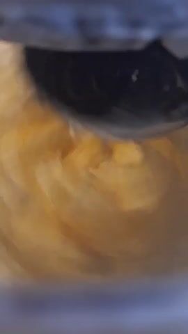 A kitchen tool with a black blade and white handle is used to cut through a thick, pale yellow substance. The substance has a smooth, creamy texture. The blade cuts through the substance, creating swirls and eddies. The scene is viewed from a high angle.
