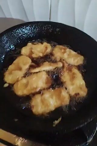 The video shows several pieces of battered food cooking in a pan of hot oil. The pieces of food appear golden brown and are arranged within the black frying pan. The oil is bubbling around the food, indicating a high cooking temperature. The background shows a white curtain and a cream colored wall. The camera remains still during the short video.