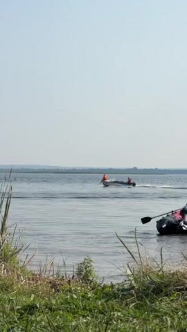 A motorboat with two people wearing orange life vests is moving across a body of water. The boat is leaving a wake behind it, indicating it is moving at a decent speed. The water appears calm with some ripples. In the foreground, tall grass and reeds grow along the bank of the water. The sky is clear and blue, with a hazy horizon in the distance. The overall scene suggests a sunny day on a lake or river.