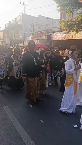 A procession is moving along a street. The procession appears to be a cultural event or a wedding. The people in the foreground are dressed in traditional attire. On the left, two individuals are wearing elaborate headdresses and ornate, colorful robes over dark clothing. In the center, a man in a black top and patterned sarong walks with a woman in a wedding dress. Behind them, other people in traditional clothing are visible, some holding parasols. The street is paved, and there are buildings lining the sides. Sunlight is casting shadows, indicating it is daytime. The overall atmosphere seems festive.