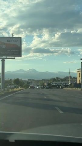 The video shows a view from a vehicle driving on a highway. The road is asphalt with white lane markings. There are several cars driving in the same direction as the camera. On the left side of the road, there is a billboard with text that is difficult to read due to the angle and resolution. Further down the road, there are buildings and trees. In the distance, there are mountains that appear to be snow-capped. The sky is partly cloudy with bright sunlight. The overall impression is a scenic drive with a backdrop of natural landscapes. The audio is music.