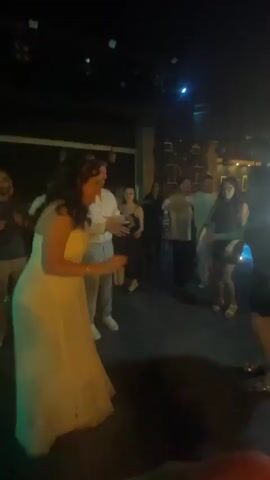 A crowd of people are dancing on a dance floor. The lighting changes between bright green and bright purple. In the foreground, a woman in a light-colored strapless dress is dancing. To her right, a woman in a black dress is also dancing. In the background, other people are dancing and socializing. The environment appears to be a nightclub or event space.
