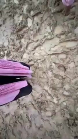 The video shows a muddy outdoor environment. In the first few seconds, someone wearing pink socks and black shoes steps onto the mud. The camera then pans to show several people wading through thick mud. Some are barefoot or wearing shorts and tank tops. The mud is brown and churned up. The video ends with a view of a crowd gathered in the mud. A canopy with colorful decorations hangs above the gathering. Loudspeakers and other structures are visible as well.