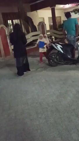 The video shows an outdoor scene at dusk.  In the foreground, a paved surface, possibly a road or sidewalk, is visible.  In the background, a building with a covered porch is illuminated.  To the left, a person wearing a black abaya stands.  In the center, two young children wearing hijabs are walking, one with a blue object in hand.  To the right, a person in a green t-shirt and jeans stands near a parked black motorcycle.  The overall impression is of a family or group of people gathered outdoors in a residential area.