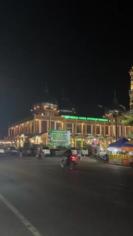 The video shows a street scene at night in front of a building with a sign that appears to be in an Asian language, possibly Indonesian. The building is illuminated and has a domed roof. There are several cars and motorcycles parked and moving on the street. Some people are visible, including individuals on motorcycles. There are also some food stalls or vendors set up on the right side of the street. The overall atmosphere appears to be that of a busy evening in a town or city.