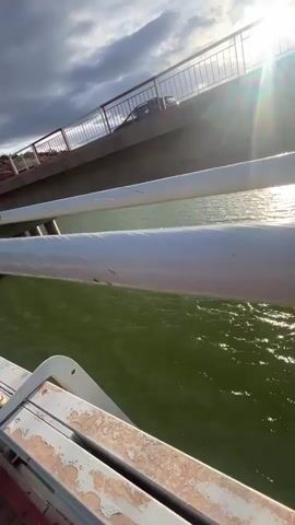 The video shows a view from a bridge with a wooden walkway and white metal railings. The bridge spans a body of water that reflects the sky. To the right of the screen is a concrete bridge with red railings. The sky above is a mix of dark, cloudy patches and areas of blue. The camera appears to be moving forward along the bridge, maintaining a low angle. On the left, suspension cables can be seen leading towards the bridge's supporting structure. The landscape in the background consists of reddish-brown hills covered with sparse vegetation.