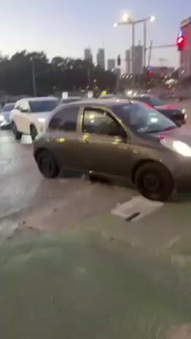 The video captures a street scene with several cars stopped at a traffic light in an urban setting. The road appears wet and reflects the ambient light. A white SUV is prominently visible, taking center stage as it slowly moves forward. Behind it, a gray Nissan Micra is partially visible. To the left of the SUV, a silver car with the license plate 638-08-325 can be seen. In the background, skyscrapers punctuate the skyline, and streetlights illuminate the intersection. The overall ambiance suggests a busy city environment at dusk or dawn.