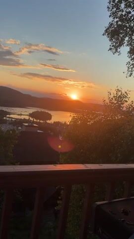 The video shows a sunset over a body of water and surrounding hills. The camera pans from left to right, offering different views of the landscape. In the foreground, a wooden deck railing is visible. The sky is filled with the warm hues of orange, pink, and yellow, with some scattered clouds. Trees and buildings are scattered throughout the landscape, with a few boats visible on the water. The overall scene is peaceful and scenic, suggesting a vacation or a quiet evening in nature.
