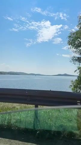 The video shows a view from a vehicle of a large body of water under a clear blue sky with scattered white clouds. There are rolling hills in the distance across the water. In the foreground, there is a metal guardrail, and below it, green grass and vegetation are visible. As the vehicle moves, the scene changes slightly, revealing a few small boats on the water, a kayaker on the shore, and a person standing in the water. A large tree is prominent on the right side of the frame at one point. The overall impression is a sunny day at a lake or reservoir.