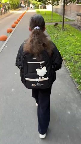 A young girl with dark hair in a ponytail walks away from the camera down a sidewalk. She is wearing a black jacket, dark pants, and white shoes. She has a black backpack on her back, adorned with a white fluffy keychain. The sidewalk is paved and borders a grassy area with trees and a brick building. Orange bollards line the edge of the sidewalk on the left. In the background, other people are walking on the sidewalk further ahead. The sun is shining, casting shadows.