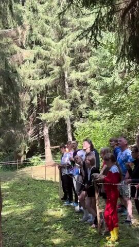 A group of people, mostly children, stand in a line on a grassy area in a forest, watching something or someone off-camera to the left. Most of them are wearing casual clothing. Some of the children are holding wooden sticks or poles. A few adults are also present at the back of the group. There is a wooden fence in front of the group, and a red and white tape barrier is visible further to the left, indicating a boundary or a designated viewing area. The background is dominated by tall evergreen trees, suggesting a wooded or forested environment. The lighting indicates it is daytime, with sunlight filtering through the trees.
