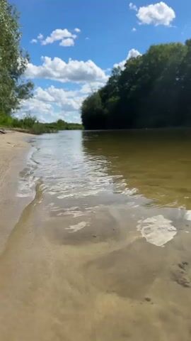 This is a close-up, low-angle video of a sandy riverbank on a sunny day. The shallow water laps gently at the shore, creating ripples and reflecting the bright blue sky dotted with fluffy white clouds. The river is calm and appears shallow near the bank, with the sandy bottom visible through the clear water. 

On the opposite bank, a dense line of lush green trees forms a natural border, their leaves a vibrant contrast to the water and sky. The scene suggests a peaceful, natural setting, likely a river or stream in a rural area. The bright sunlight and blue sky indicate it is daytime, probably late morning or early afternoon.

The location is identified as Borisoglebsk, Russia. The overall mood is serene and tranquil, evoking a sense of calm and natural beauty. There is no visible text in the media. The focus is on the natural elements: sand, water, trees, sky, and clouds, creating a visually appealing and calming composition. The gentle movement of the water and the play of light on its surface are prominent details.