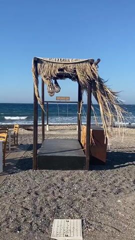 A beachside scene is captured under a clear blue sky. In the foreground, a rustic structure made of dark wood and thatched roofing stands on a pebble beach. The structure has a raised platform in the center, and on either side, there are what appear to be seating areas or decorative elements covered in straw. Suspended from the thatched roof are three glass-like objects encased in netting. A wooden sign with the word "DRUNKFISH" is visible in the center of the structure, just above a set of metal swings. Beyond the structure, the ocean stretches out with moderate waves rolling towards the shore. To the left, a portion of a wooden chair is visible. The scene is calm and appears to be a seaside establishment.
