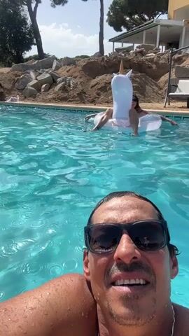 A man in sunglasses takes a selfie in a swimming pool, with a woman on an inflatable unicorn float in the background. The pool is surrounded by a concrete deck and a rocky, landscaped hillside with trees and a building on top. The water is clear and blue, with ripples indicating movement. The man is smiling and looking at the camera. The woman on the float is also looking towards the camera and smiling. The sky is visible in the background, appearing clear and bright.