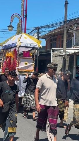 The video shows a group of men carrying a decorated structure through a street on a sunny day. The structure is a canopy-like construction adorned with white fabric, golden decorations, and red and gold patterned fabric. It is supported by bamboo poles that the men carry on their shoulders. The men are dressed in a variety of clothing, including black t-shirts, traditional patterned wraps, and headbands. Some are smiling and laughing, while others appear more serious. The street is lined with buildings of varying heights and architectural styles, and there are power lines visible overhead. The overall impression is of a procession or ceremony taking place in a residential area.