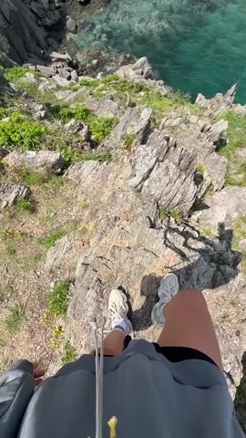 The video shows a person standing on a rocky cliff overlooking a bay. The water is clear and turquoise in color. There are other people visible on the cliff. The sky is partly cloudy.