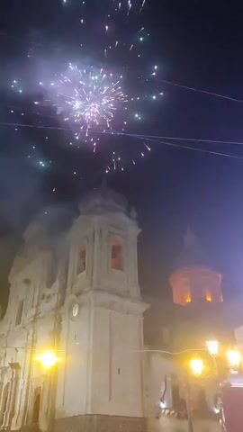 The video shows a nighttime outdoor scene in front of a church. Fireworks are being set off in the sky, creating bursts of light and smoke. In the foreground, a crowd of people is gathered, many of them looking up at the fireworks. There are also streetlights and decorative lights visible, illuminating the scene. The church has a bell tower and ornate architecture. The overall atmosphere appears to be celebratory.