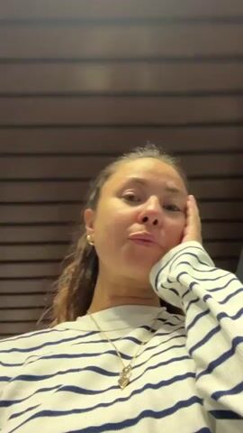 A woman is filmed from a low angle looking up at the camera. She has long brown hair and is wearing a striped sweater and a necklace. She is resting her head on her hand and looking towards the camera with a neutral expression. Above her, there are three circular lights in a rectangular fixture, and the ceiling appears to be made of wood planks. The lighting is bright, and the scene is indoors.