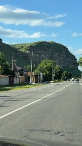 This video captures a view from a vehicle driving along a paved road with white lane markings. The scene is set in a rural or semi-rural area, with a distinctive, large, layered rock formation dominating the background. The rock formation is covered in green vegetation, indicating a temperate climate and likely springtime or summer. A bright blue sky with scattered white, fluffy clouds suggests fair weather.

Along the side of the road, there are utility poles with wires stretching across the frame. A few buildings are visible, including what appears to be a small shop or market with signage. A yellow bus and other vehicles are driving on the road, indicating it's an active thoroughfare. A pedestrian crossing sign and a bus stop shelter are also present, suggesting a settled area.

The time of day appears to be daytime, likely late morning or afternoon, given the bright sunlight casting shadows. The overall mood is peaceful and scenic, highlighting the natural beauty of the landscape combined with signs of human habitation and activity. The location is identified as Ordzhonikidzevsky, Russia. There is some visible text on a building sign, which appears to read "МИНИМАРКЕТ" (MINIMARKET).