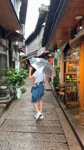 The video shows a young girl with blonde hair wearing a white t-shirt, a denim skirt, and white sneakers. She is walking down a narrow, cobblestone street while holding a clear umbrella over her head. The street is lined with traditional buildings with wooden awnings and balconies. There are plants and potted greenery along the sides of the street, and a sign with Chinese characters is visible on the right side of the frame. The girl turns to face the camera, holding a drink in her hand and touching her hair. It appears to be raining, as evidenced by the umbrella and the wet pavement.