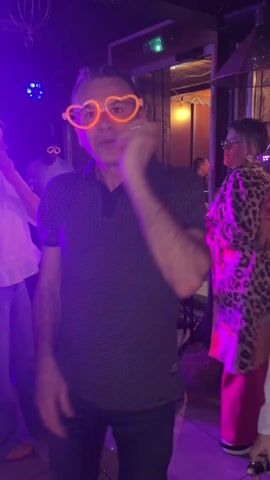 A smiling middle-aged man wearing orange heart-shaped glasses dances at a party. He is pointing directly at the camera and making various hand gestures. The room is illuminated with colorful disco lights and other partygoers can be seen in the background. The decor suggests it may be a pub or event space. He wears a dark polo shirt and dark pants.